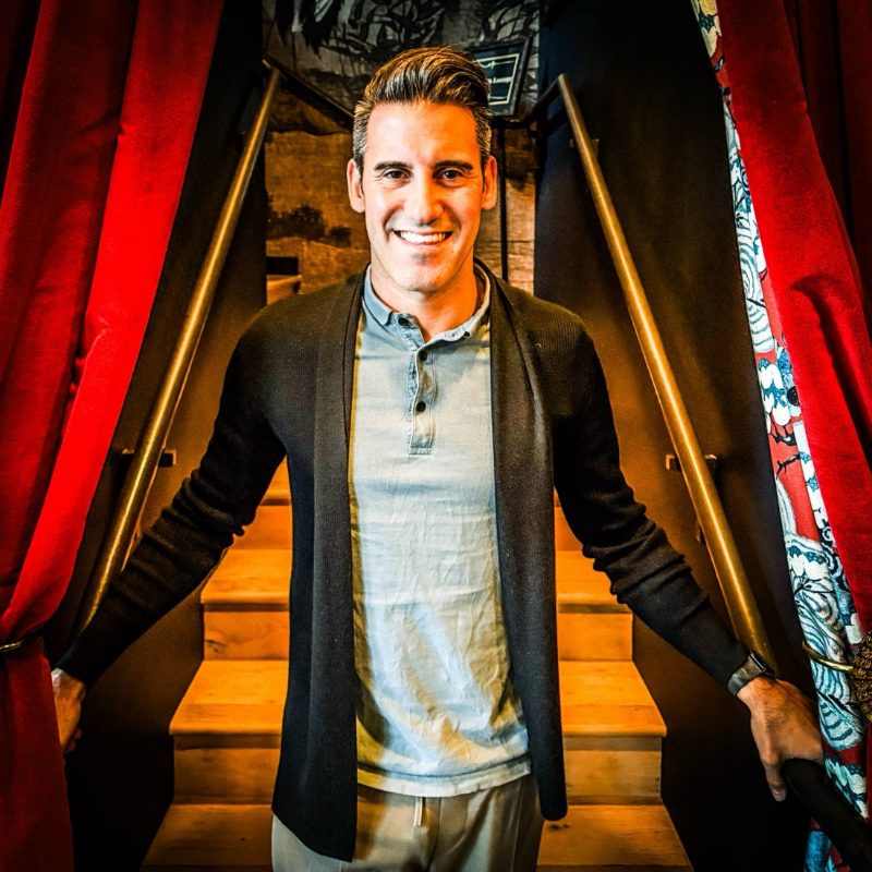 Joel smiling at the bottom of a staircase framed by red velvet curtains
